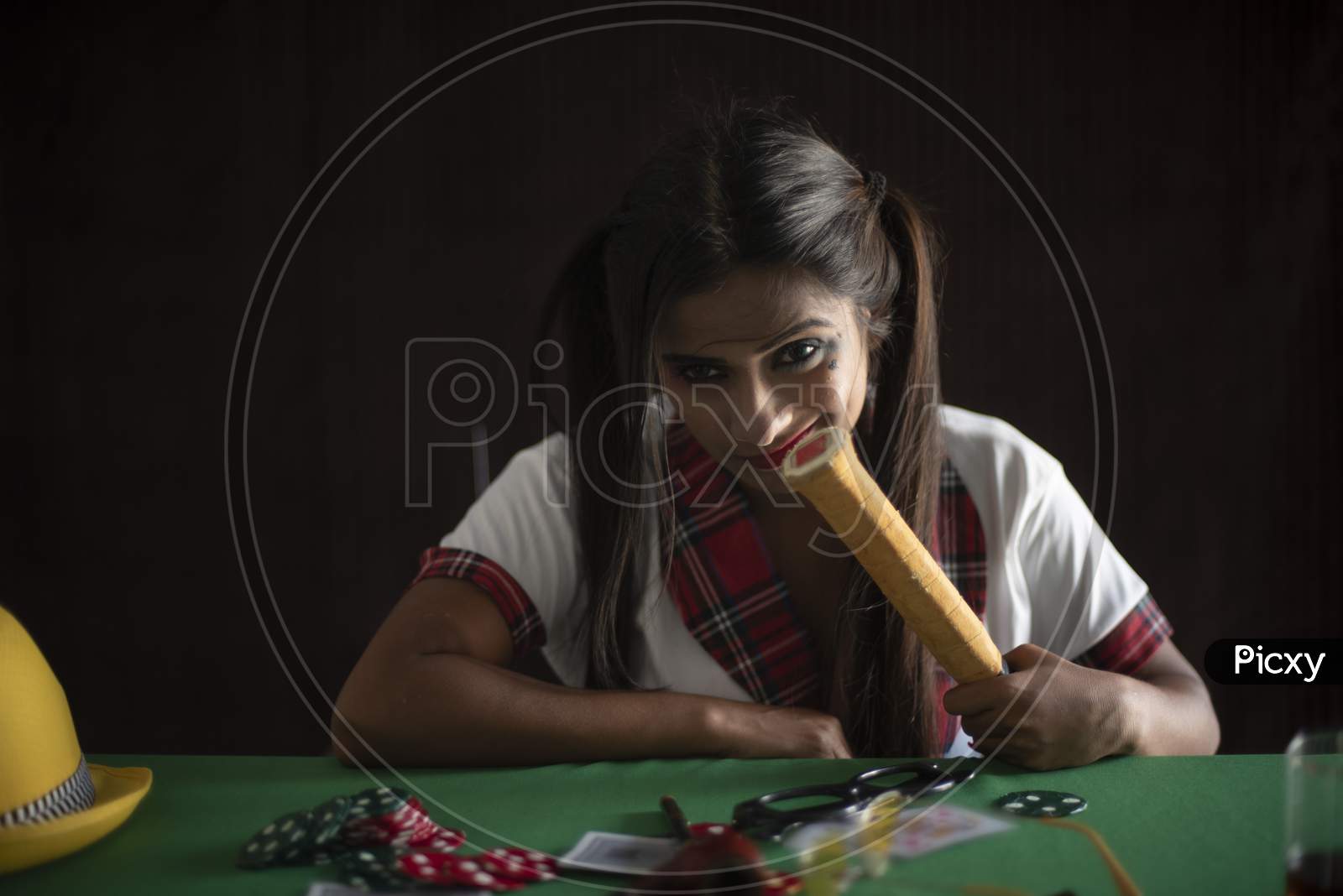 Young Indian Bengali brunette woman in school uniform with a tennis racket sitting on a casino poker table in brown textured copy space studio background. Indian lifestyle and fashion.