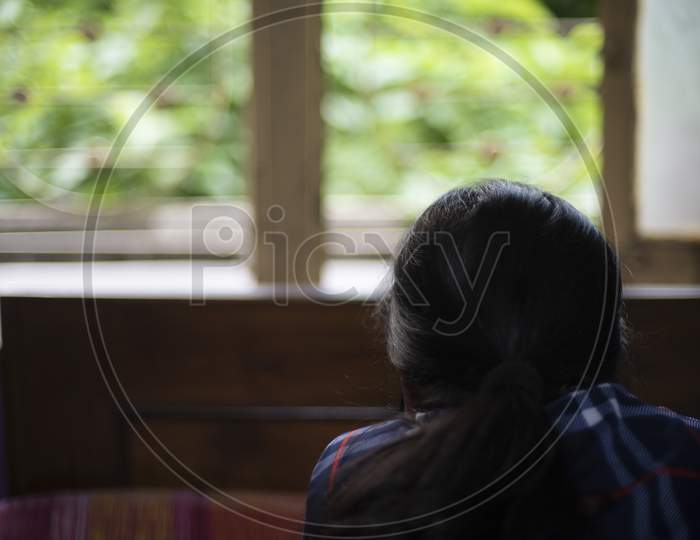 Back portrait of an Indian young woman wearing corona preventive mask in home isolation in front of a window. Indian lifestyle and disease.