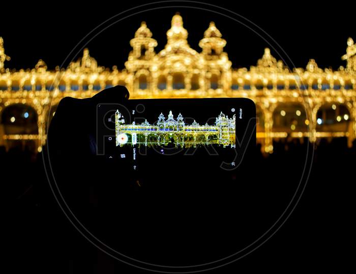A mobile phone is placed in a hand the image of Mysore Palace is on the screen and the Mysore Palace is in the background completely blurred.