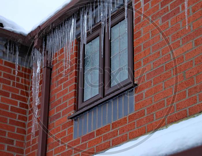 Long Icicles Hang From The Gutter Of A House. The Roof Is Covered In Snow And It'S Still Snowing