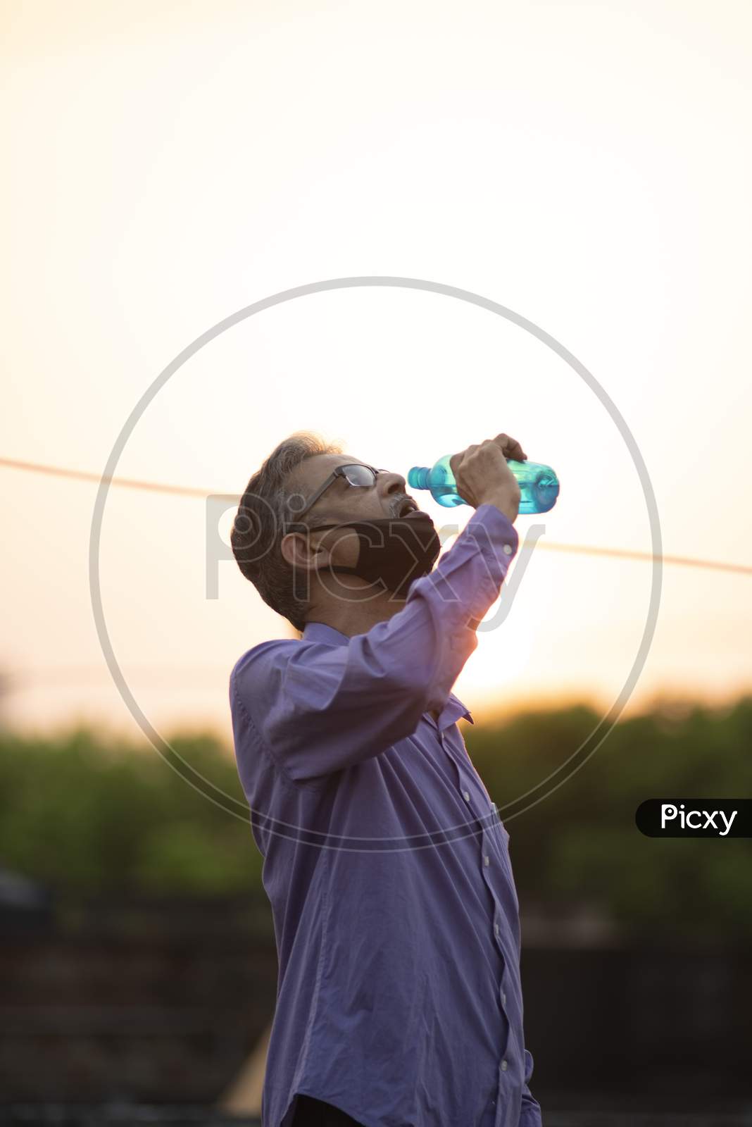 Portrait of an Indian old man with corona preventive mask drinking water from a bottle on a rooftop  in home isolation.Indian lifestyle, disease and home quarantine.
