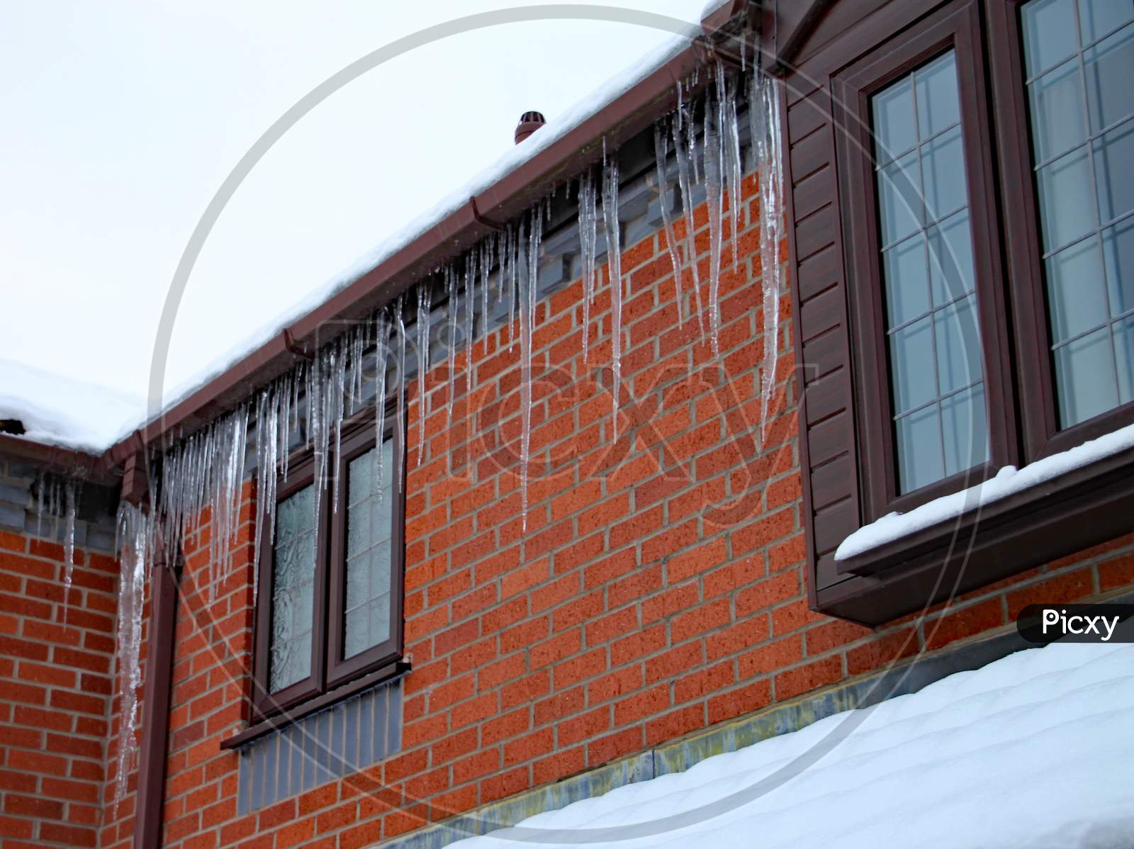 Long Icicles Hang From The Gutter Of A House. The Roof Is Covered In Snow And It'S Still Snowing