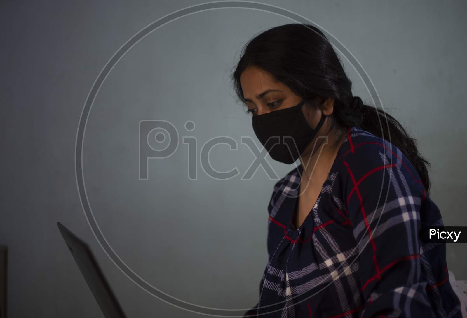 Portrait of an Indian young woman with Corona preventive mask sitting at home with a laptop home isolation due to covid - 19. Indian lifestyle and disease.