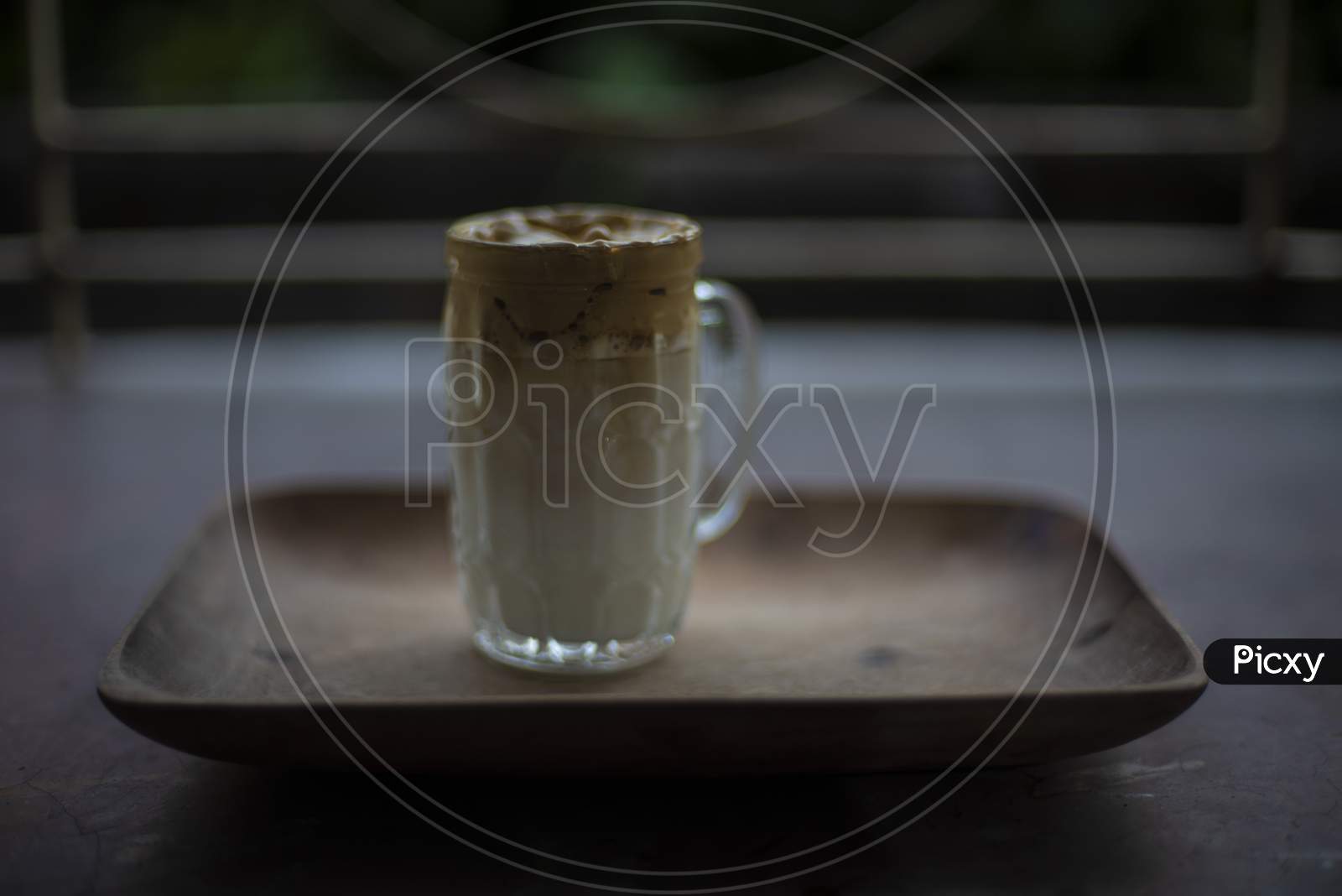 A glass of Dalgona coffee on a wooden tray kept in front of a window. Refreshment and drink