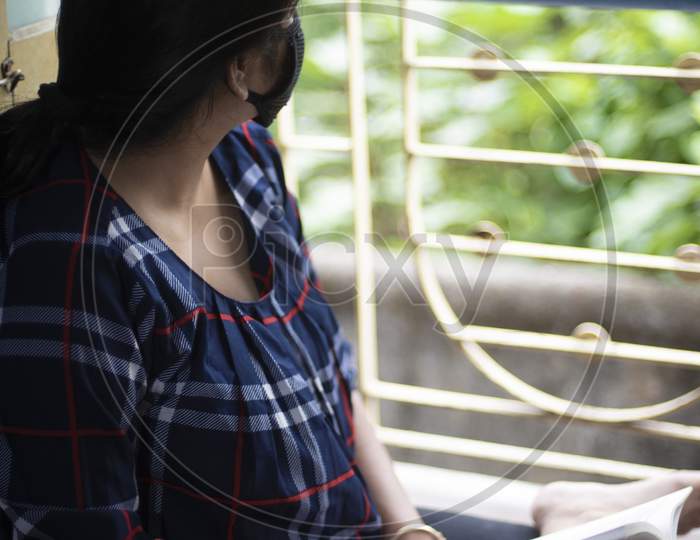 Portrait of an Indian young woman wearing corona preventive mask reading book in home isolation in front of a window. Indian lifestyle and disease.
