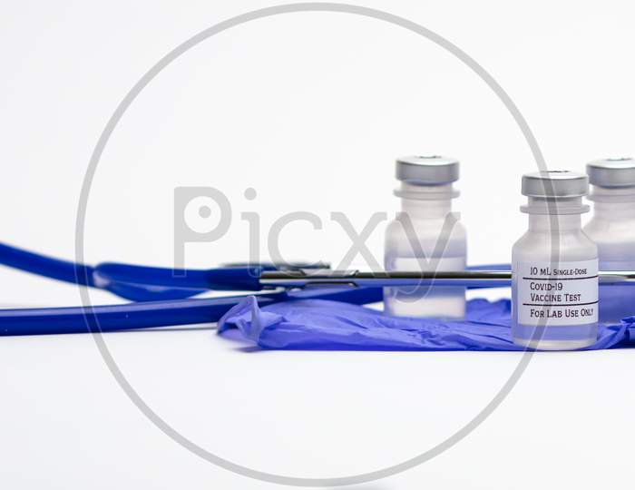 Three Covid-19 Test Vaccine Vials On Top Of Blue Medical Gloves And Surrounded By A Blue Stethoscope.
