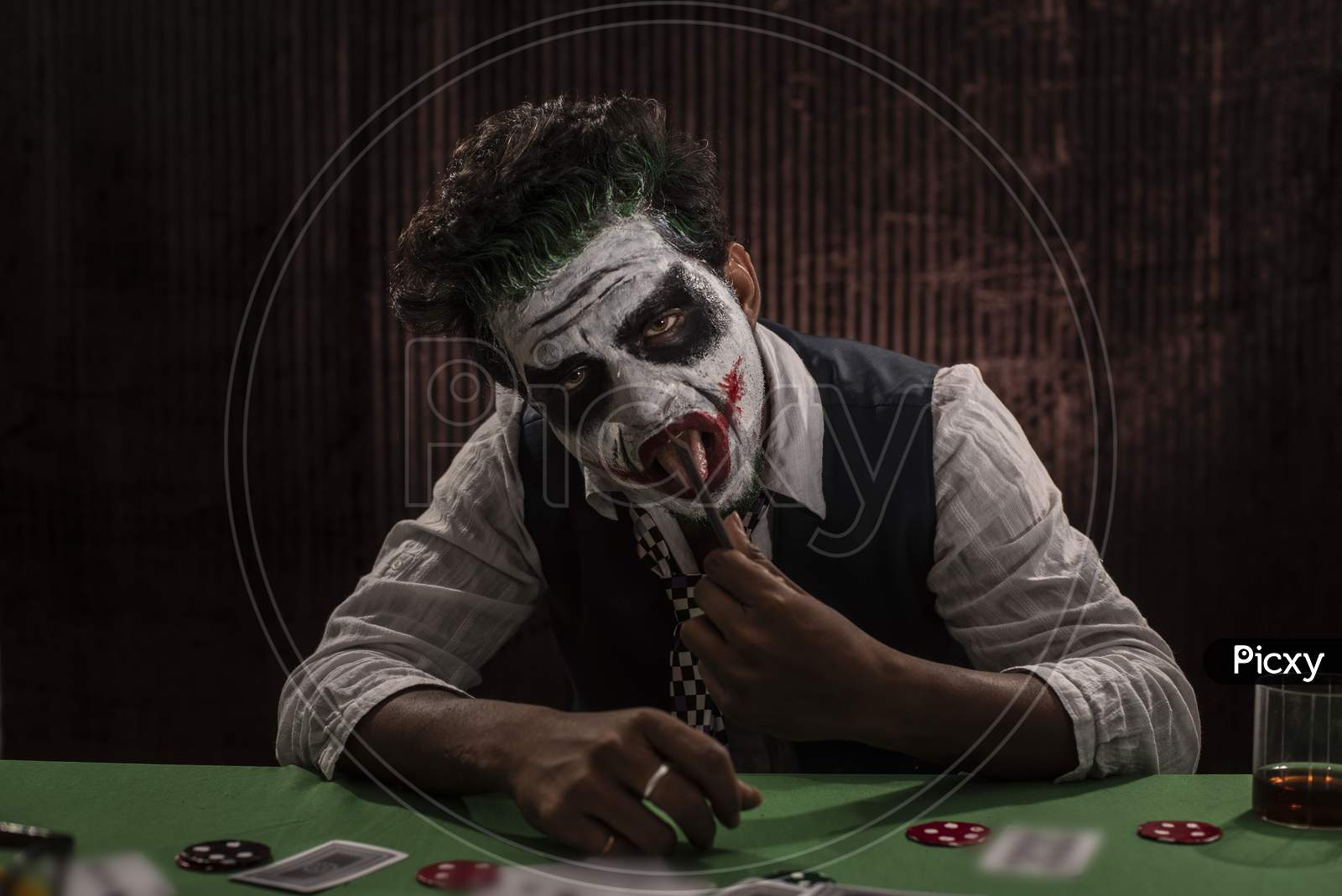 Portrait of an Indian man in Halloween Joker costume showing scary facial expression in front of a casino poker table. Cosplay photography.