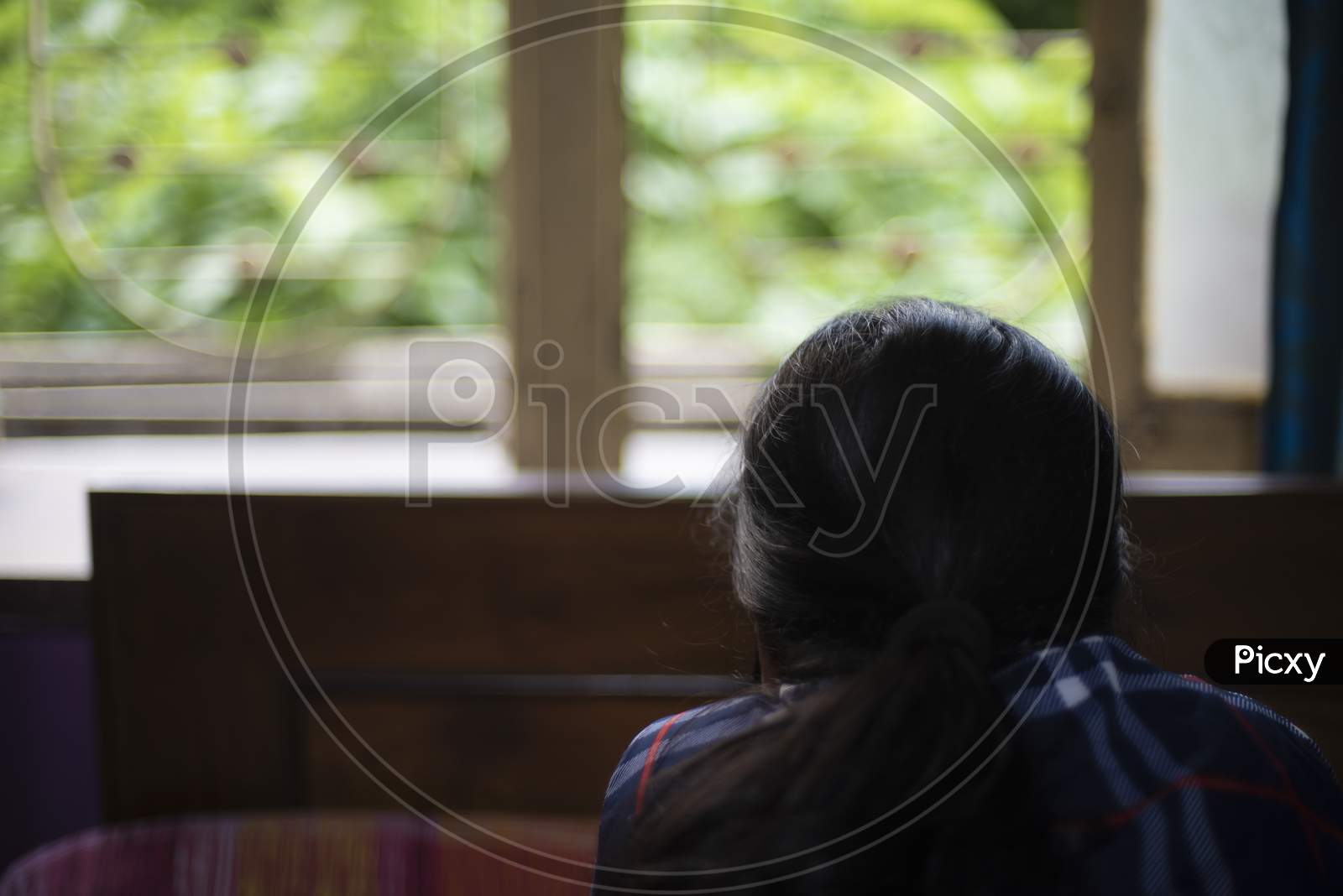 Back portrait of an Indian young woman wearing corona preventive mask in home isolation in front of a window. Indian lifestyle and disease.