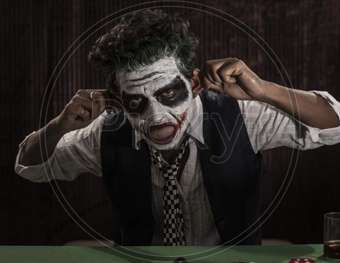 Portrait of an Indian man in Halloween Joker costume showing scary facial expression in front of a casino poker table. Cosplay photography.