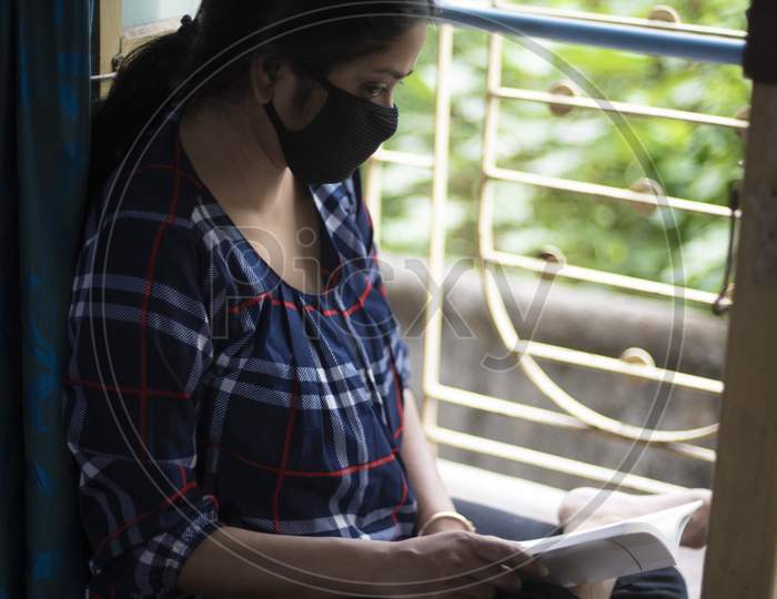 Portrait of an Indian young woman wearing corona preventive mask reading book in home isolation in front of a window. Indian lifestyle and disease.