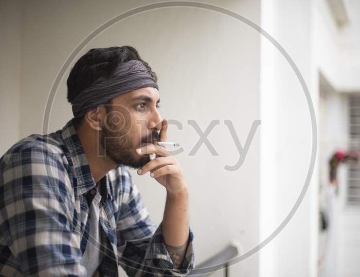 Indian Bengali handsome beard macho guy/man/male in sun glasses and head band smoking cigarette standing on balcony in white urban background. Indian lifestyle.
