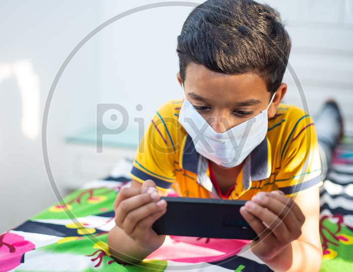 Young Boy Wearing Mask Lying Down On A Bed In A Relaxed Pose, Playing At His Smart Phone. Kid Staying Home During The Quarantine. Social Distance, Learning Online Education, Covid-19 Lock Down.