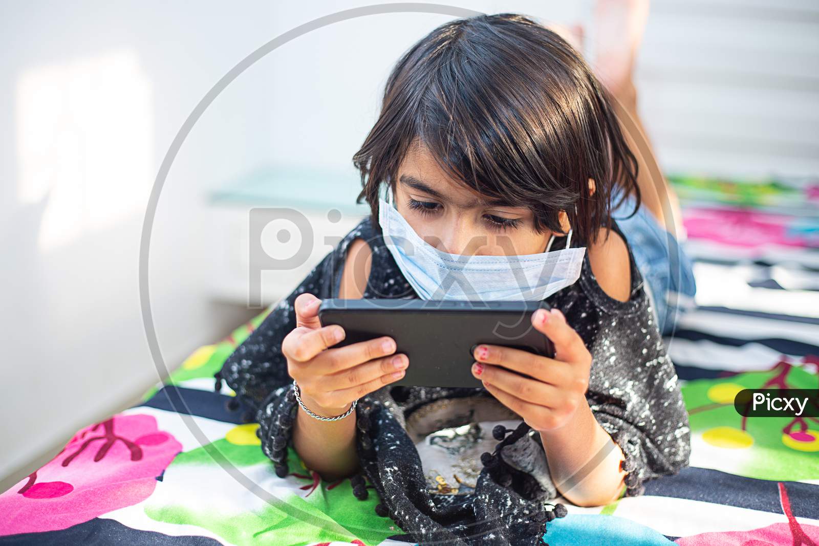 Young Preschool Girl Wearing Mask Lying Down On A Bed In A Relaxed Pose, Playing At Her Smart Phone. Staying Home During The Quarantine. Social Distance, Learning Online Education, Covid-19 Lock Down.