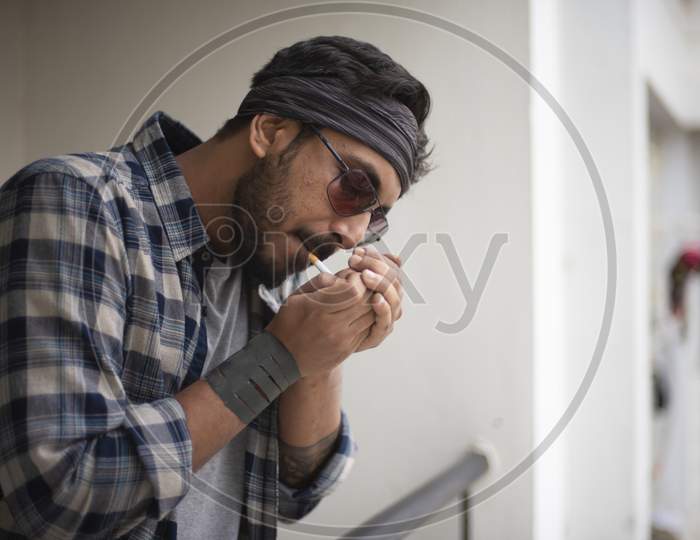 Indian Bengali handsome beard macho guy/man/male in sun glasses and head band smoking cigarette standing on balcony in white urban background. Indian lifestyle.
