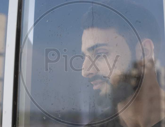 Blurred portrait of young brunette Indian Kashmiri man in casual tee shirt looking outside in front of a glass window with droplets of water and dewdrops. lifestyle and fashion.