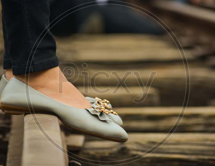 Foot (Feets) Of A Girls Standing On A Railway Track Wearing Grey Coloured Bellies With A Golden Flower On It. Old Fashioned Railway Tracks.