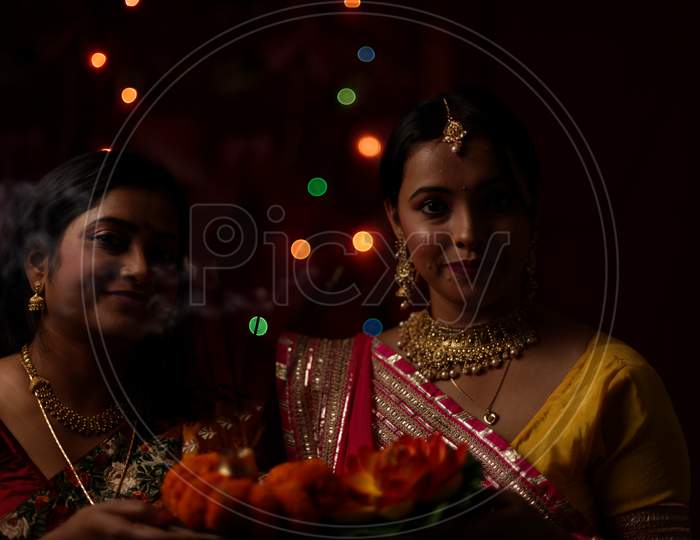 Two beautiful Indian Bengali women in Indian traditional dress celebrating Diwali with incense sticks and flowers in light bokeh background on Diwali evening. Indian lifestyle and Diwali celebration