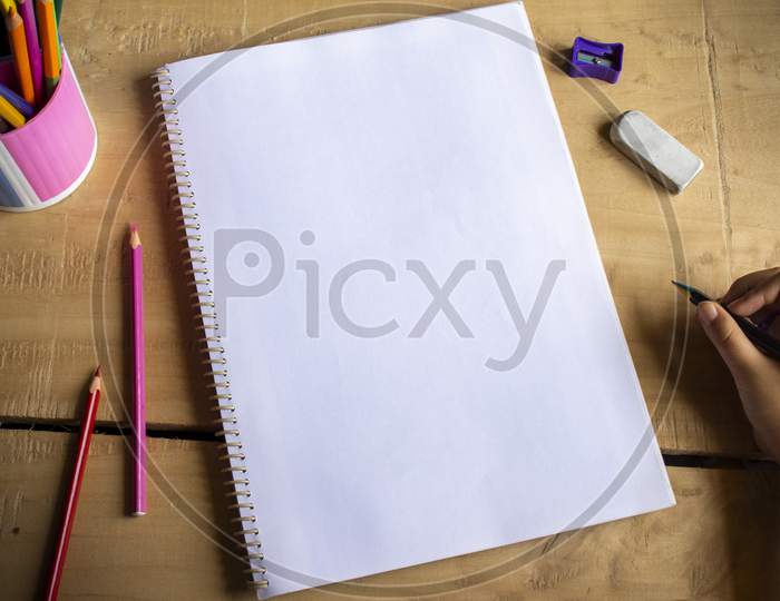 Top View Of An Empty White Notebook Space For Text, Color Pencils, Eraser, Sharpener And Human Hand Holding Pencil On Wooden Table