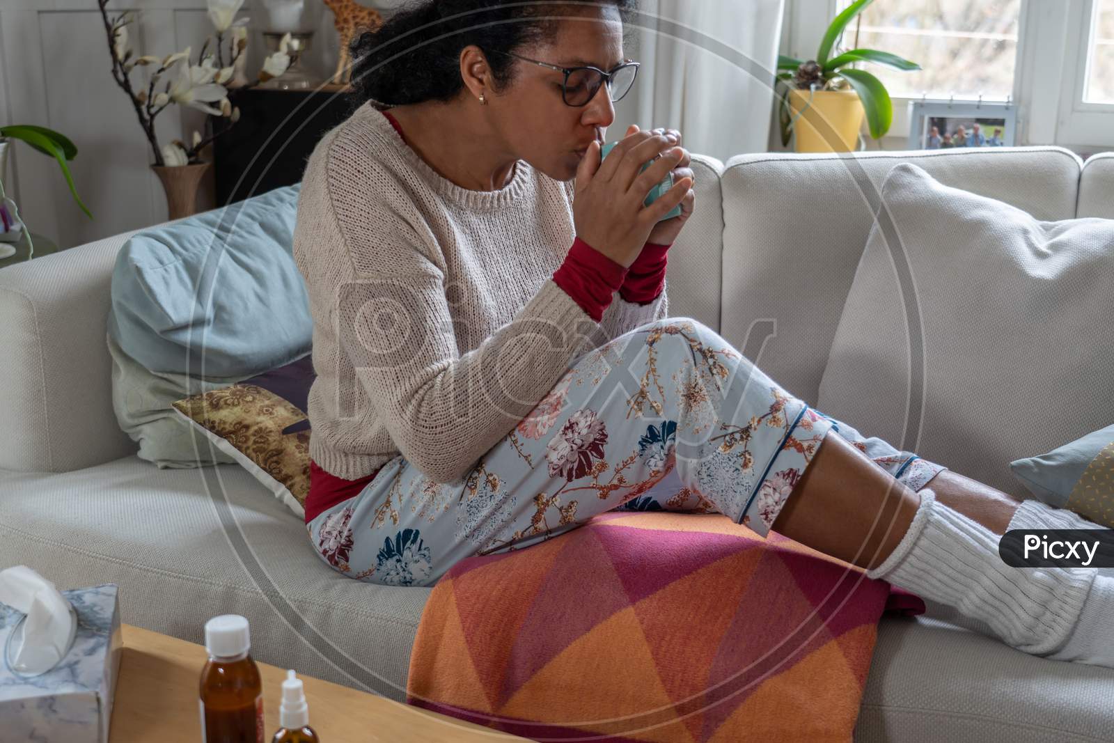 Woman having a cold, sitting on white couch in the living room while drinking tea. Medicine and tissue paper on table.
