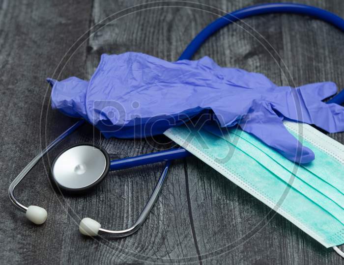 A Pair Of Blue Rubber Medical Gloves, Surgical Mask And Blue Stethoscope On A Wooden Table.