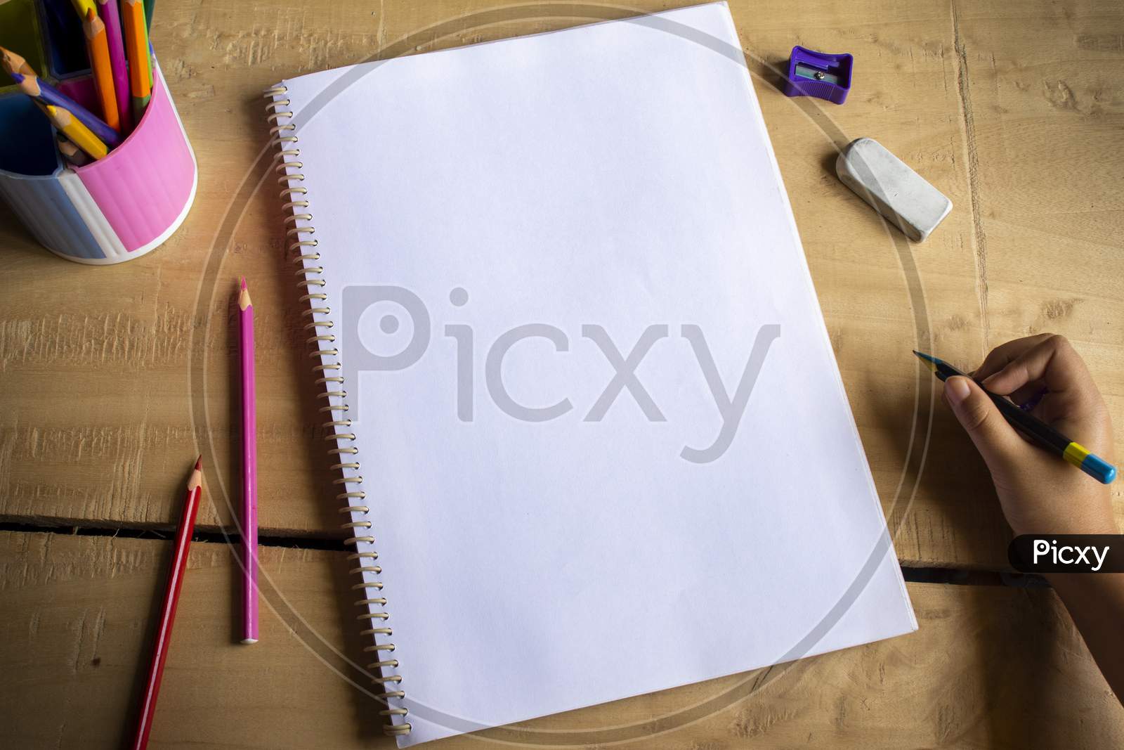Top View Of An Empty White Notebook Space For Text, Color Pencils, Eraser, Sharpener And Human Hand Holding Pencil On Wooden Table