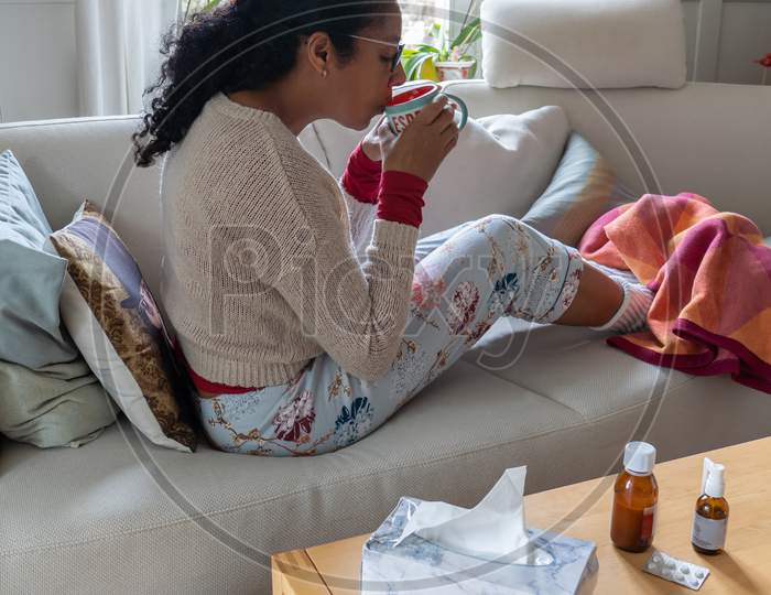 Woman having a cold, sitting on white couch in the living room while drinking tea. Medicine and tissue paper on table.