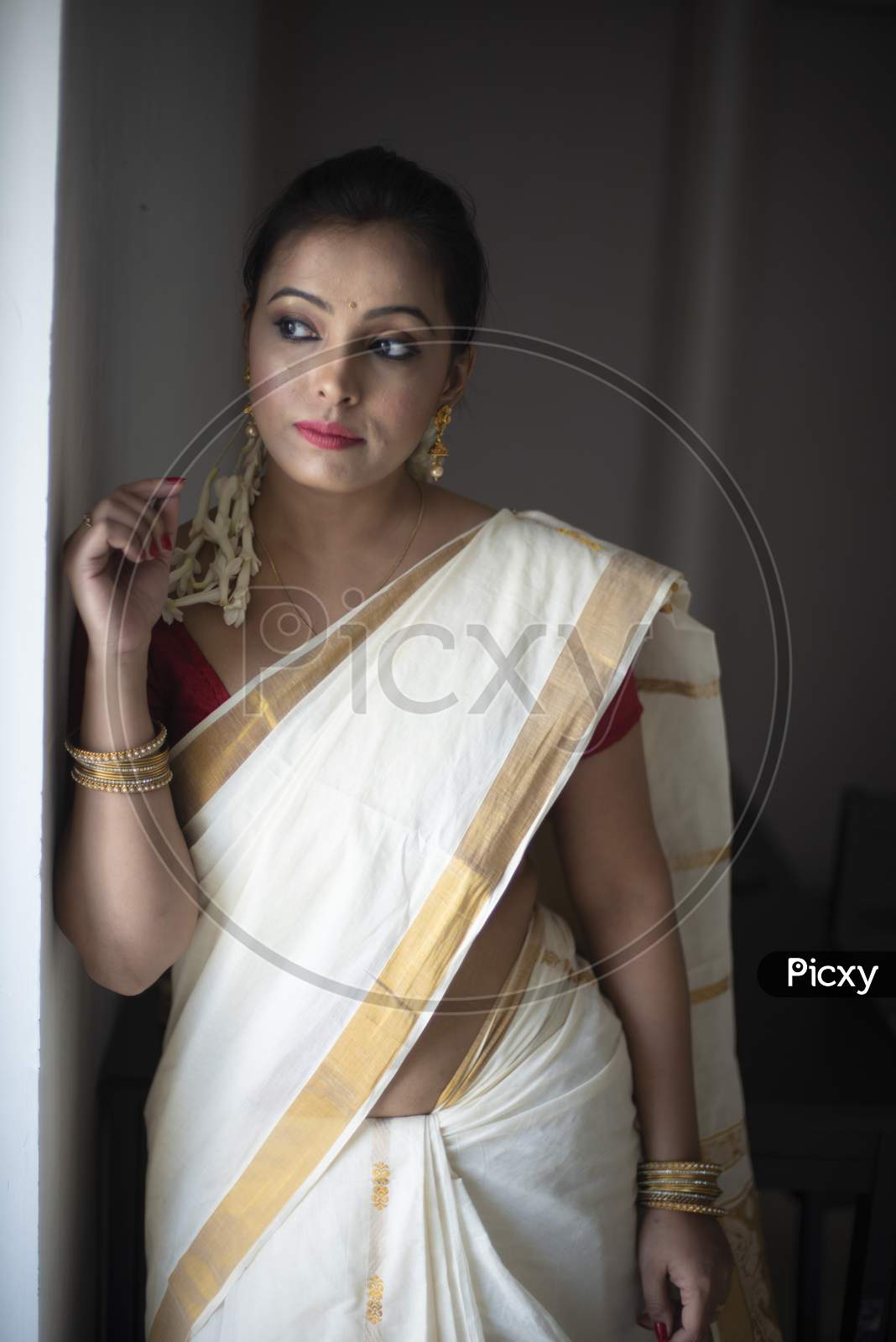 An young and attractive Indian woman in white traditional sari and red blouse and flowers is smiling while Standing at a Balcony and Posing   for the celebration of Onam/Pongal. Indian lifestyle.
