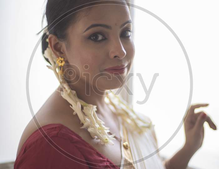 An young and attractive Indian woman in white traditional sari and red blouse and flowers is smiling while standing in front of a glass window for the celebration of Onam/Pongal. Indian lifestyle.
