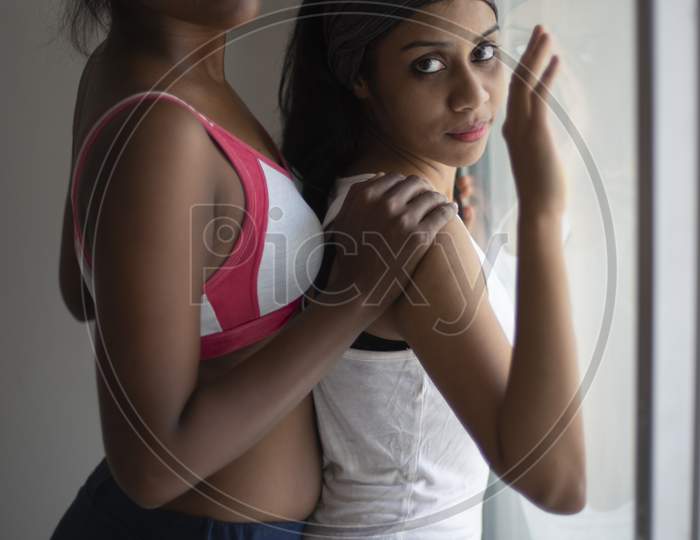 A beautiful and young Indian Bengali lesbian couple in sports inner/underwear are interacting in a intimate way in front of a glass window in white background. Dark and fare model, Indian lifestyle