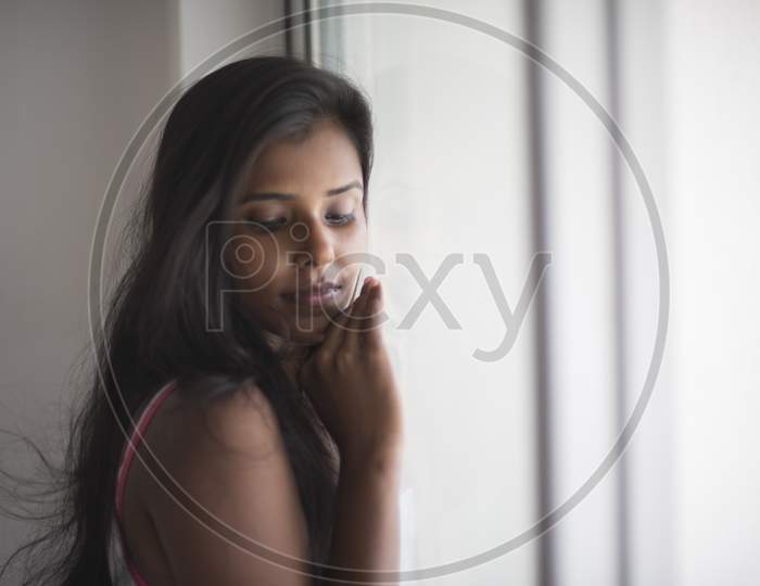 Portrait of an attractive young brunette dark skinned Indian Bengali girl in western sportswear standing At Window Glass  in white urban background. Indian lifestyle