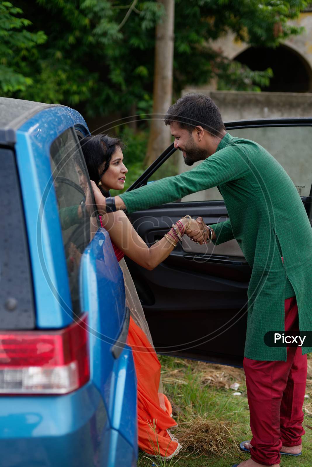 Young and attractive Indian Bengali brunette woman trying to get out of a blue car wearing Indian traditional ethnic cloths while her man is by opening door. Indian lifestyle and fashion