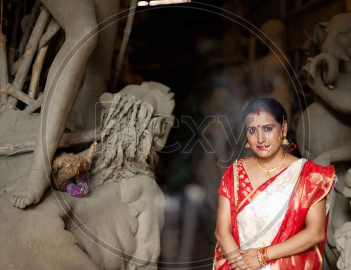 Portrait of an young and beautiful Indian Bengali brunette woman in red and white traditional ethnic sari in front of the clay idol of Hindu Bengali goddess Durga. Indian culture, religion and fashion