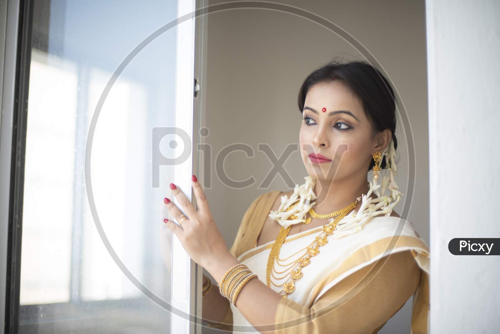 An young and attractive Indian woman in white traditional sari and red blouse and flowers is standing in a balcony for the celebration of Onam/Pongal. Indian lifestyle.