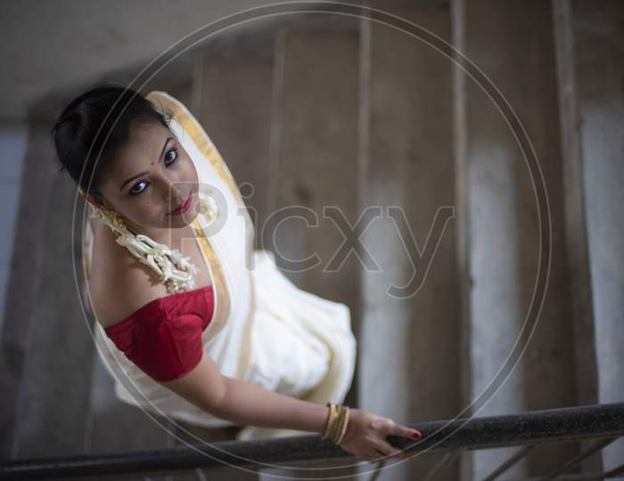An young and attractive Indian woman in white traditional sari and red blouse and flowers is Walking on Steps or Stairs for the celebration of Onam/Pongal. Indian lifestyle.