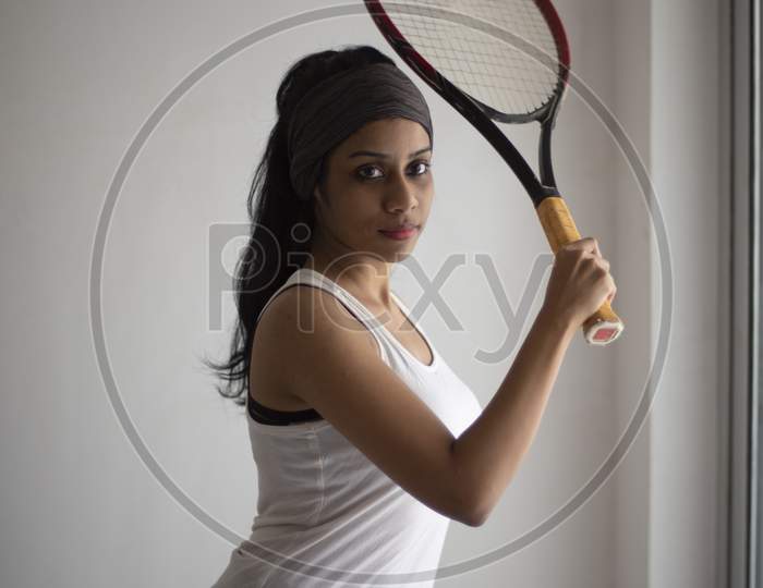 Portrait of an attractive young brunette Indian Bengali girl in western sportswear and a tennis bat in a balcony in white background. Indian lifestyle
