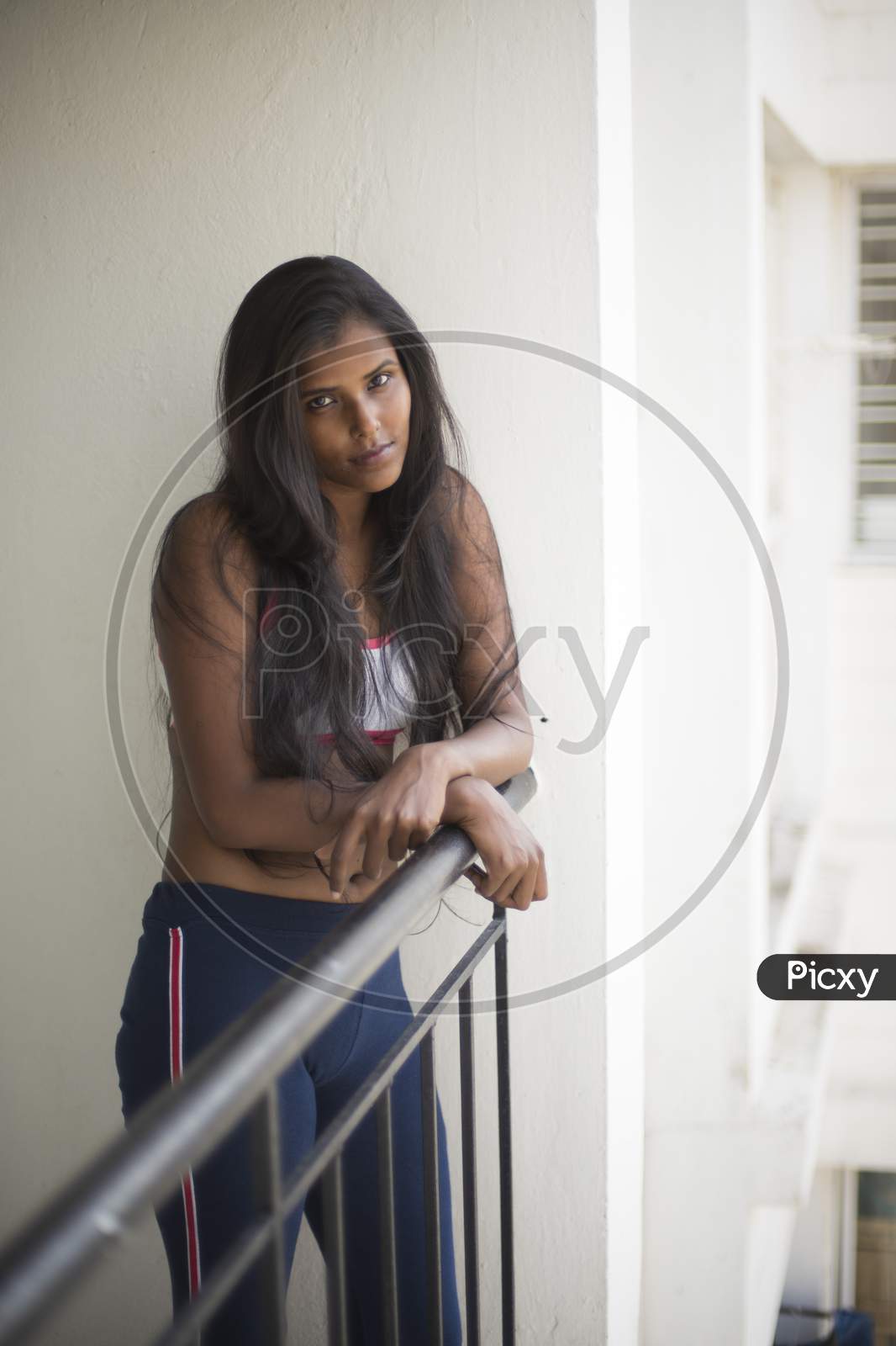 Portrait of an attractive young brunette dark skinned Indian Bengali girl in western sportswear standing on a balcony in white urban background. Indian lifestyle