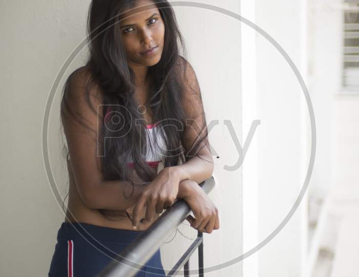Portrait of an attractive young brunette dark skinned Indian Bengali girl in western sportswear standing on a balcony in white urban background. Indian lifestyle