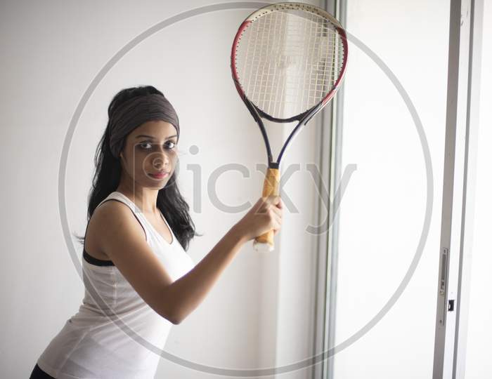 Portrait of an attractive young brunette Indian Bengali girl in western sportswear and a tennis bat in a balcony in white background. Indian lifestyle