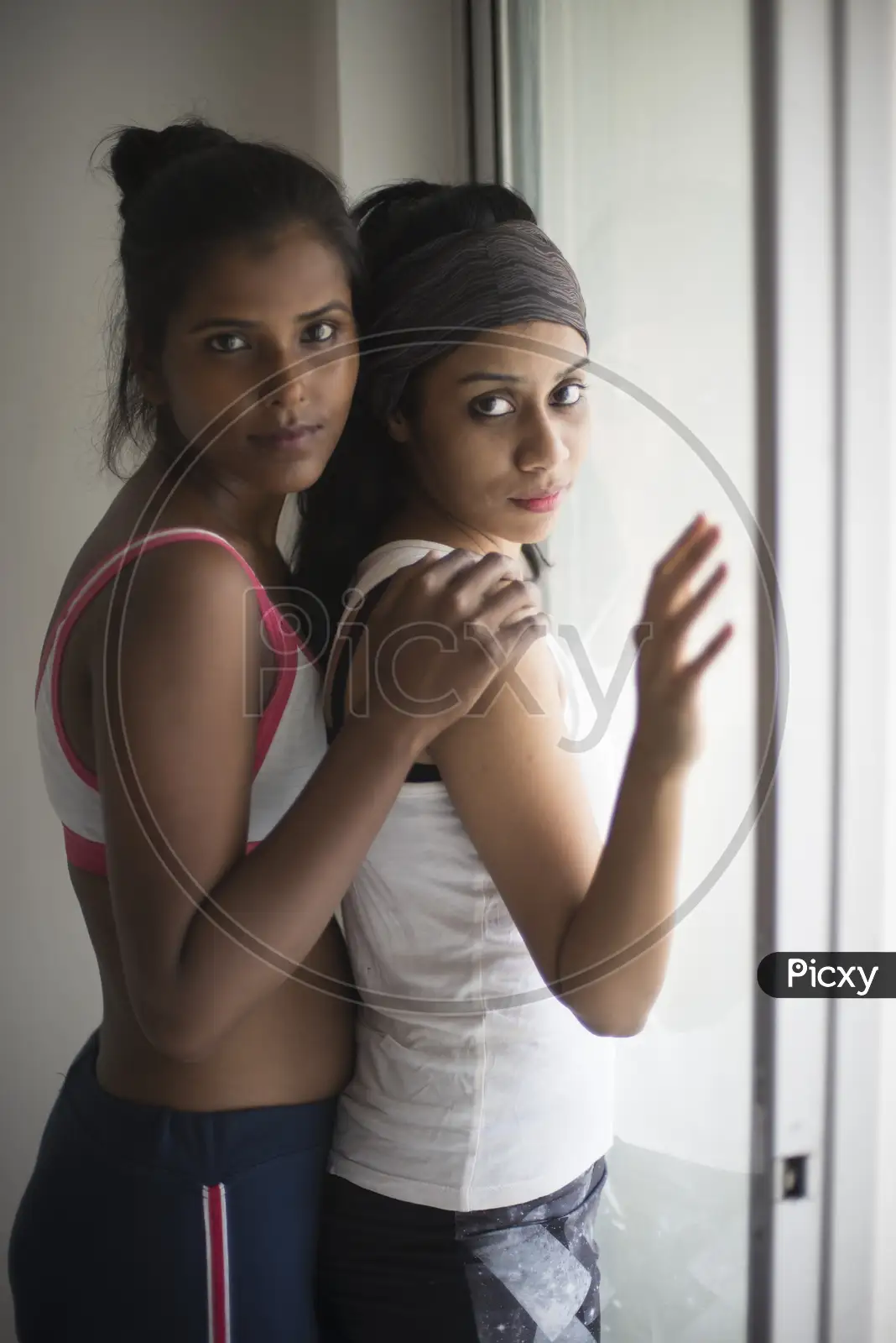 Image of A beautiful and young Indian Bengali lesbian couple in sports  inner/underwear are interacting in a intimate way in front of a glass  window in white background. Dark and fare model,