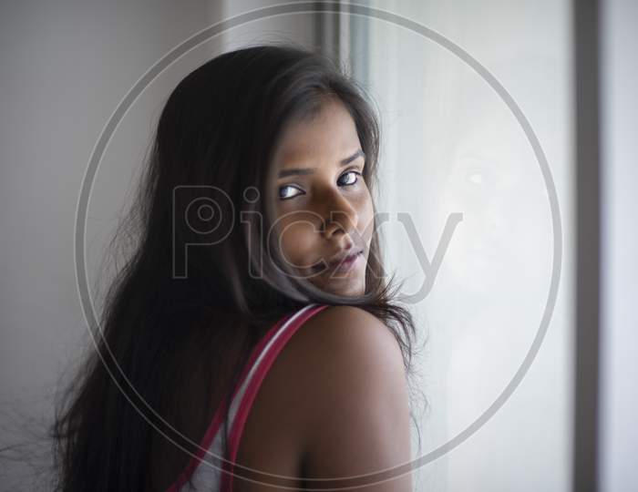 Portrait of an attractive young brunette dark skinned Indian Bengali girl in western sportswear standing at Window Glass  in white urban background. Indian lifestyle