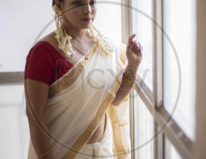 An young and attractive Indian woman in white traditional sari and red blouse and flowers is smiling while standing in front of a glass window for the celebration of Onam/Pongal. Indian lifestyle.