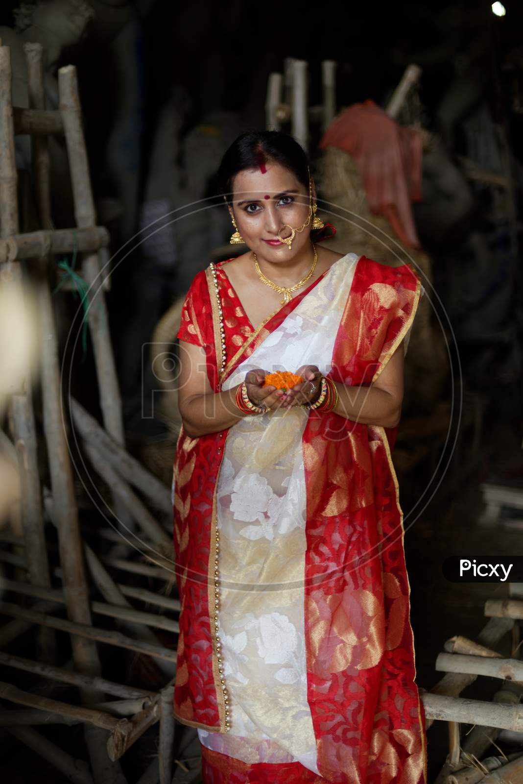 Portrait of an young and beautiful Indian Bengali brunette woman in red and white traditional ethnic sari in front of the clay idol of Hindu Bengali goddess Durga. Indian culture, religion and fashion
