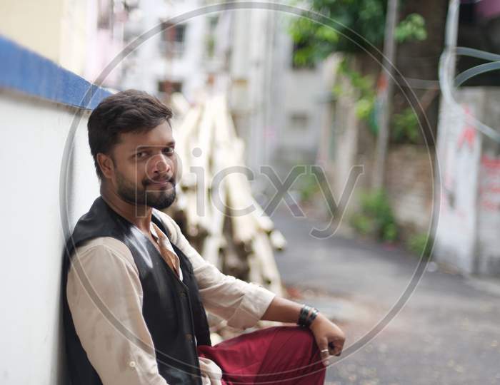 portrait of young and handsome Indian Bengali brunette beard man posing on street leaning on a wall wearing Indian ethnic vibrant jacket, shirt and red pant. Indian lifestyle and fashion