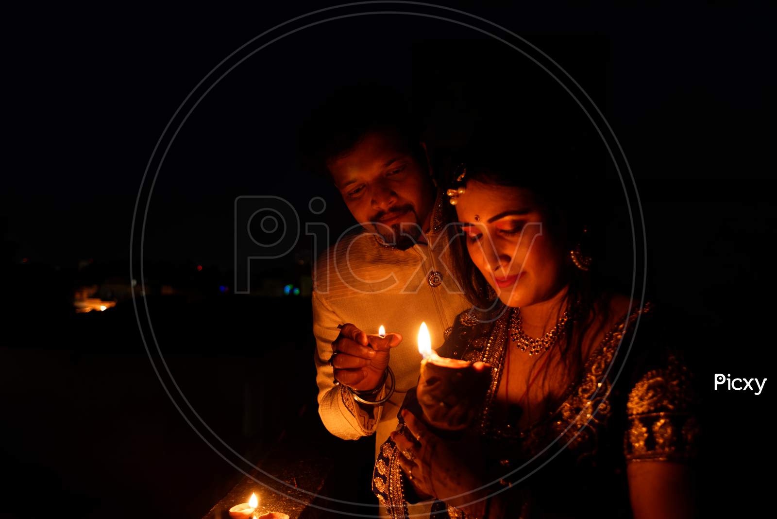 Young and beautiful Indian Gujarati couple in Indian traditional dress lightening Diwali diya/lamps on the terrace in darkness on Diwali evening. Indian lifestyle and Diwali celebration