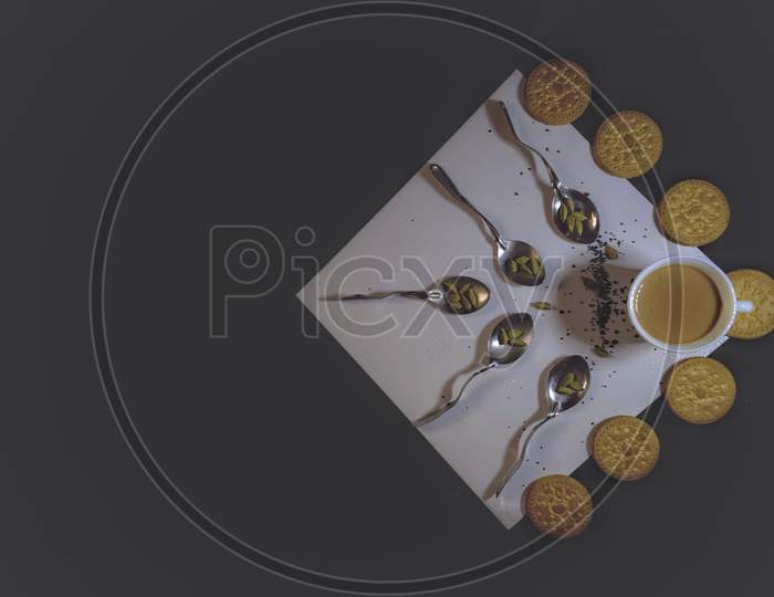 A cup of tea with creatively placed spoons and biscuits on a white plate over a black background.