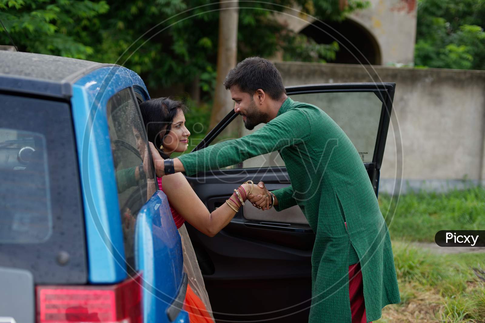 Young and attractive Indian Bengali brunette woman trying to get out of a blue car wearing Indian traditional ethnic cloths while her man is by opening door. Indian lifestyle and fashion