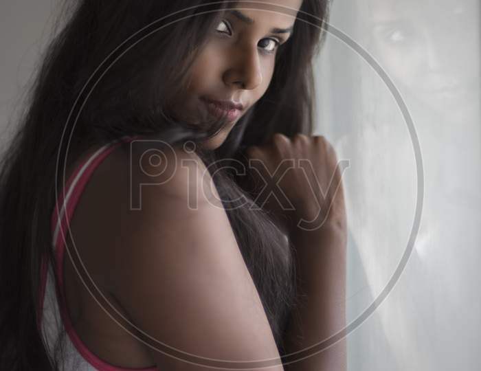 Portrait of an attractive young brunette dark skinned Indian Bengali girl in western sportswear standing At Window Glass  in white urban background. Indian lifestyle