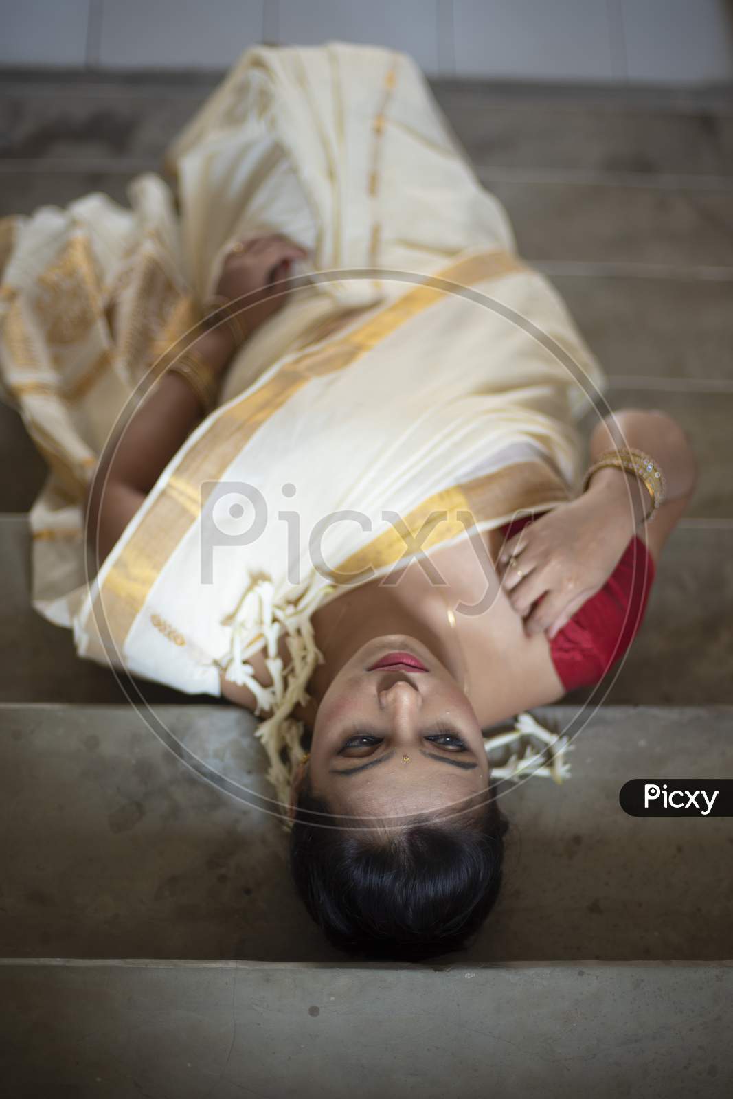 An young and attractive Indian woman in white traditional sari and red blouse and flowers is smiling while Lying On Steps  for the celebration of Onam/Pongal. Indian lifestyle.