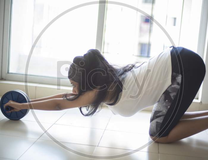 Side posture of an Indian brunette girl in sportswear performing exercise with ab roller in front of glass window in a white background. Indian lifestyle