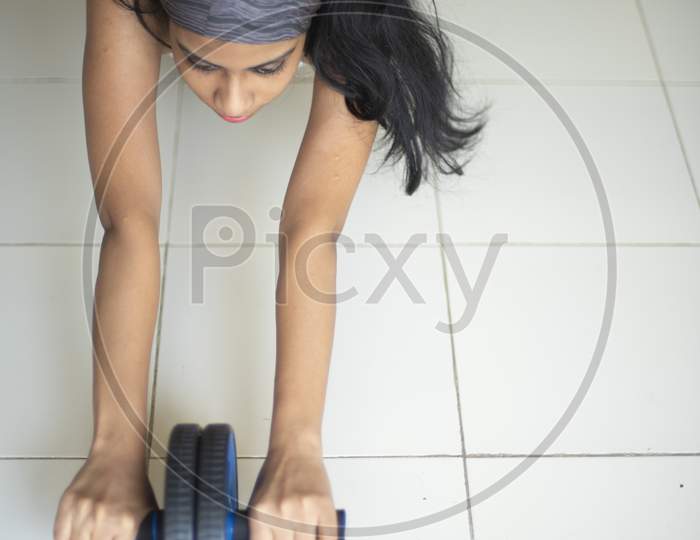 posture of an Indian brunette girl in sportswear performing exercise with ab roller in front of glass window in a white background. Indian lifestyle